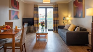 A modern living room with a sofa, a dining table with fruit, a TV, and a large window with curtains. - PREMIER SUITES Bristol Redcliffe (Bristol)