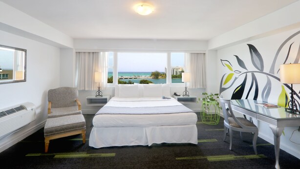 A hotel room with a large bed, a desk, a chair, a view of the ocean, and a window with curtains. - Hotel Miramar (San Juan)