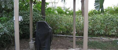 A covered outdoor seating area with a grill and a table, surrounded by greenery.