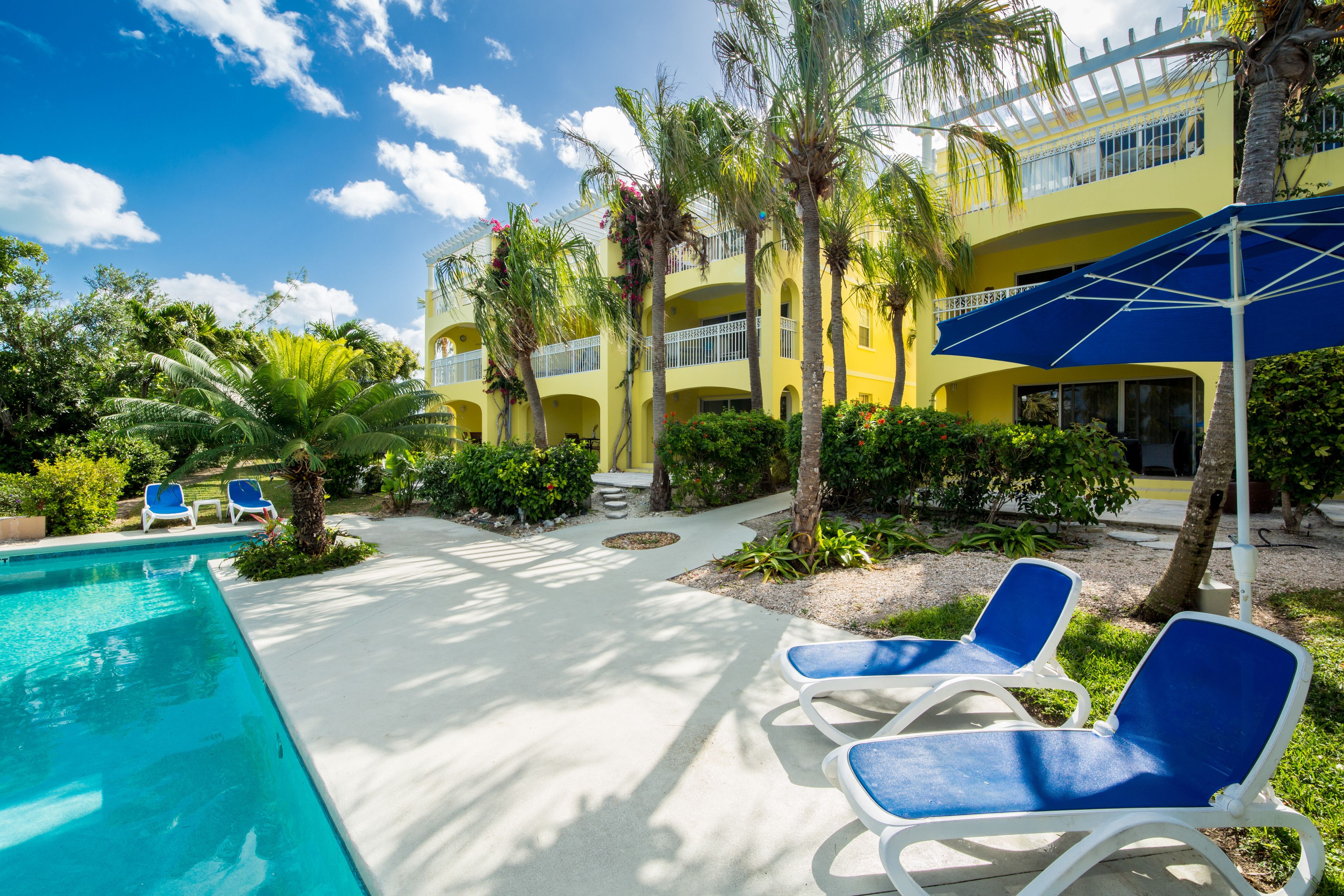 A pool area with lounge chairs, a clear sky, and a yellow building with balconies.