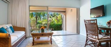 A hotel room with a sofa, a dining table with chairs, a glass coffee table, a TV mounted on the wall, and a sliding glass door leading to a balcony with a view of greenery.