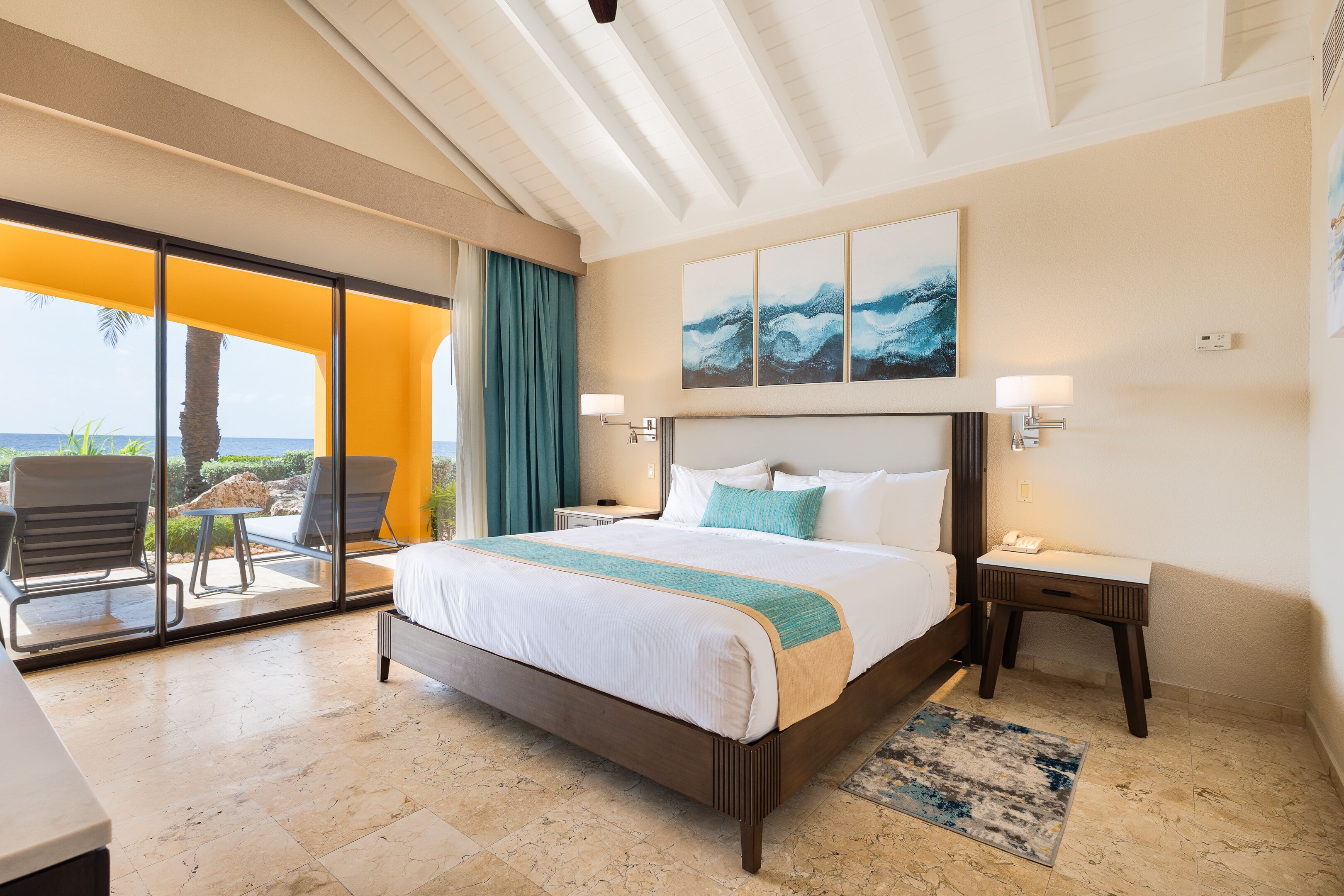 A hotel room with a large bed, a desk, a chair, a view of the ocean, and a balcony with chairs.