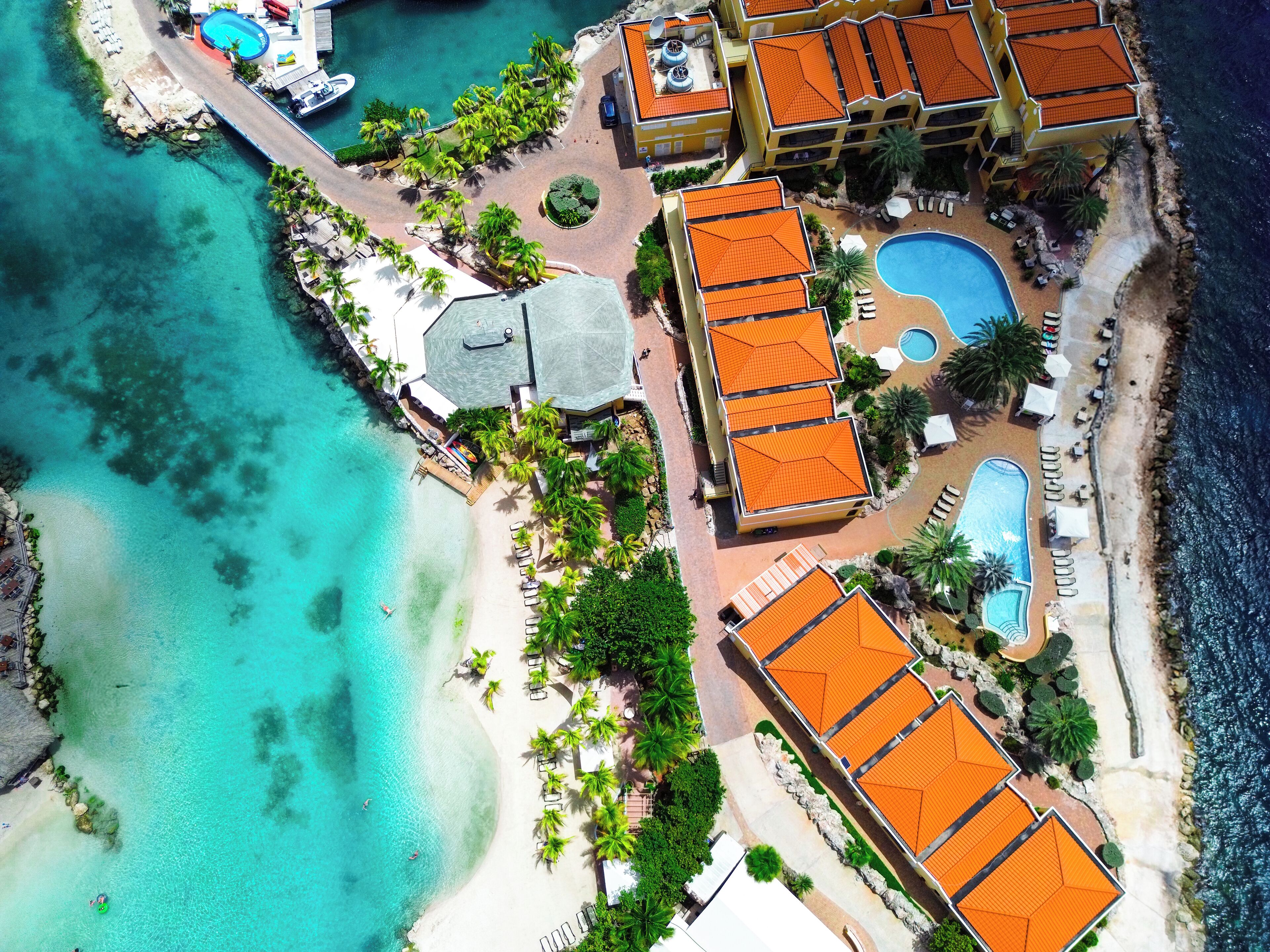 An aerial view of a resort with multiple swimming pools, a beach, and a variety of buildings with orange roofs.