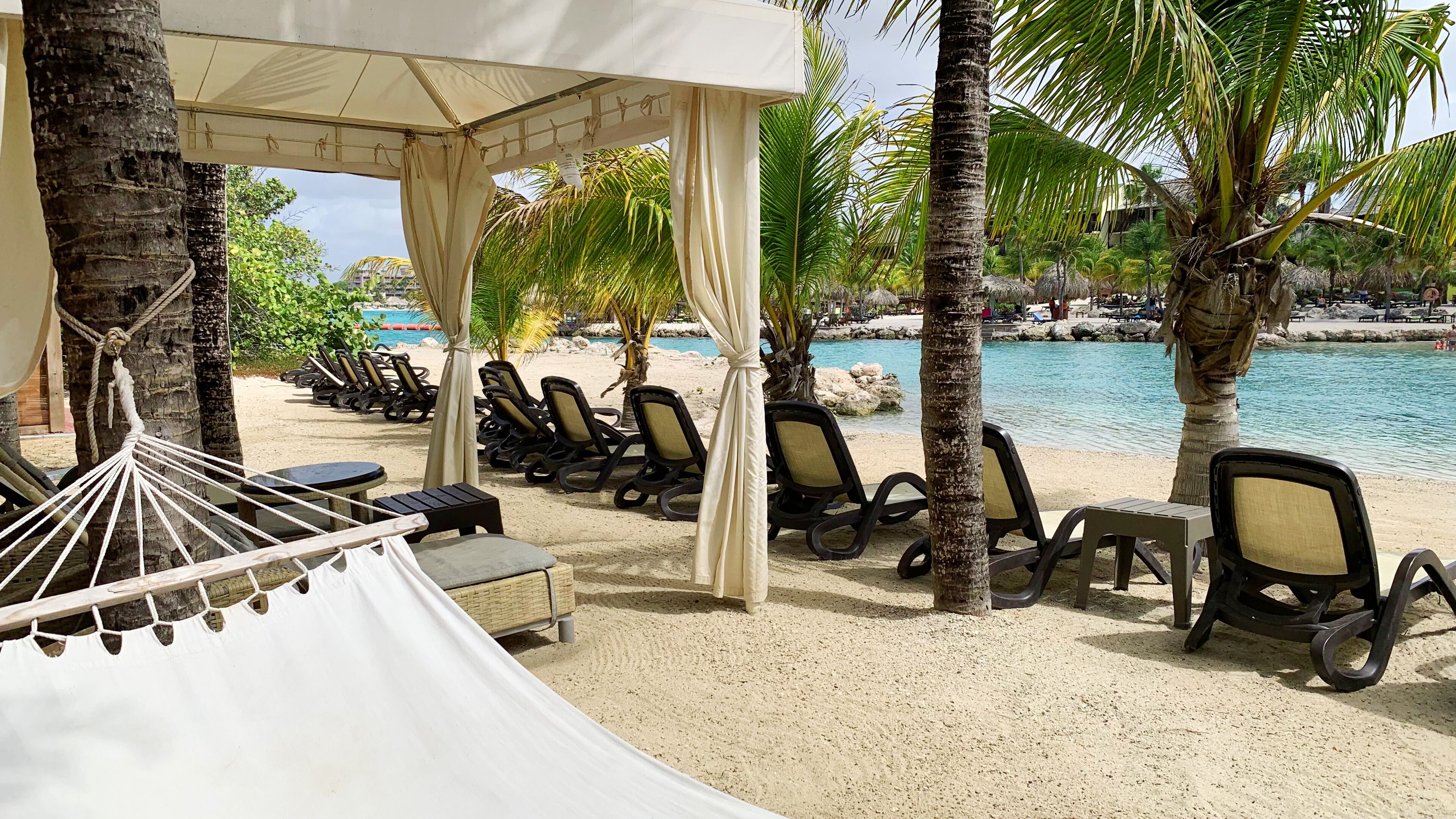 A beachside lounge area with lounge chairs, a hammock, and a canopy structure.
