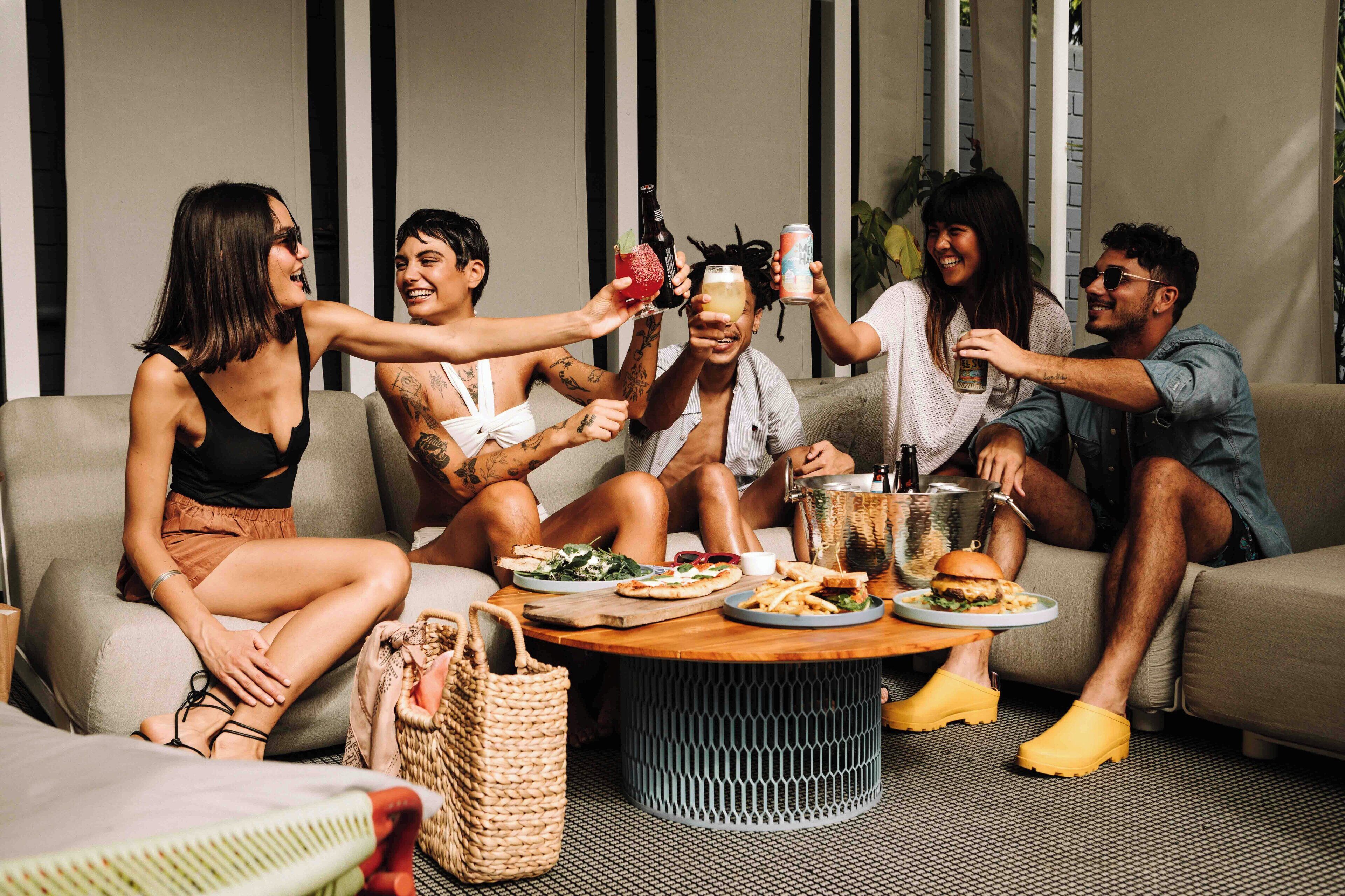 Un groupe de personnes est assis sur un canapé, levant leurs verres pour trinquer, avec de la nourriture et un panier sur une table devant eux.
