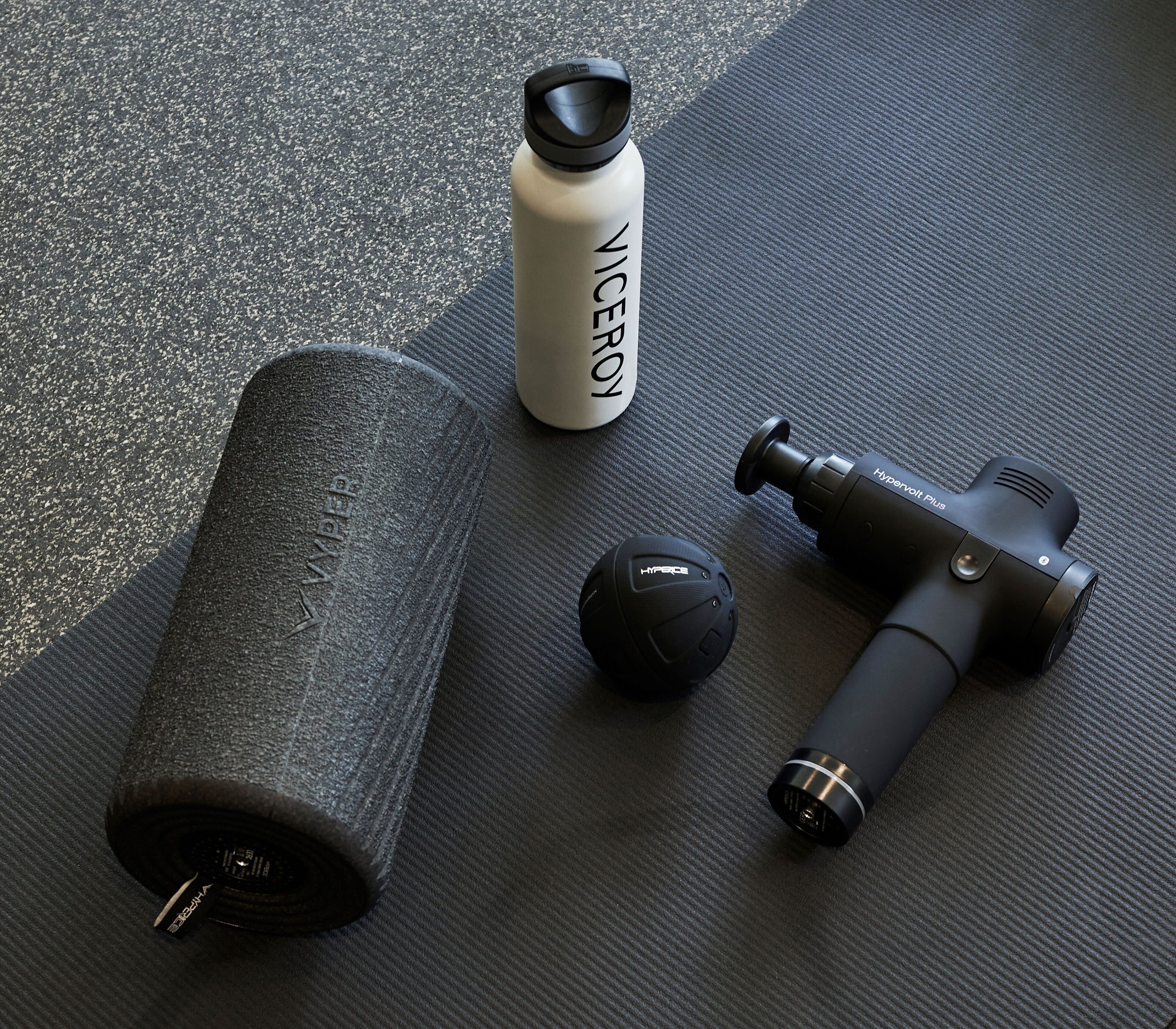 Una colchoneta de yoga, una botella de agua y un masajeador muscular sobre un suelo con textura.
