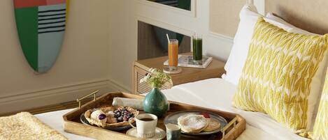 A hotel room with a bed, a tray with breakfast items, a wall-mounted surfboard, and a window with curtains.