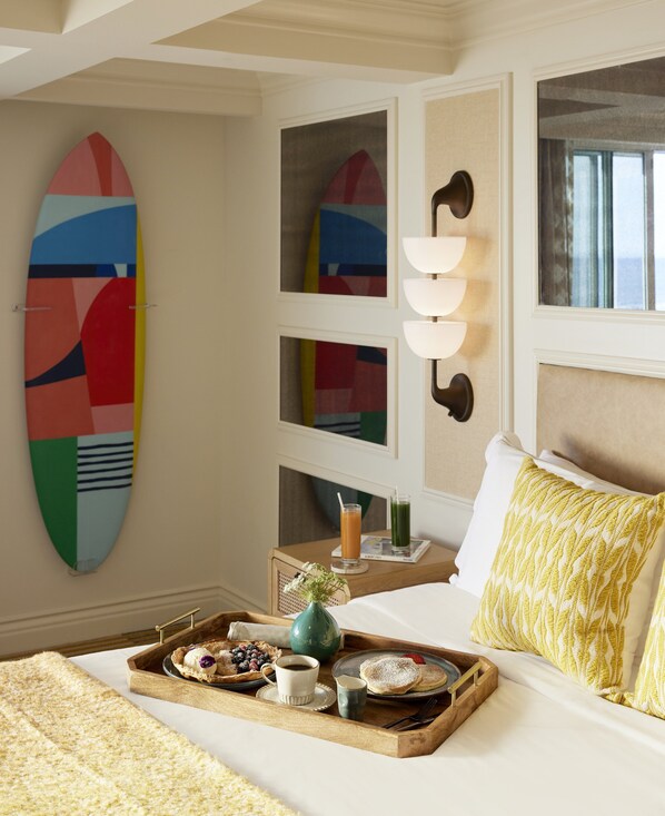 A hotel room with a bed, a tray with breakfast items, a wall-mounted surfboard, and a window with curtains. - Viceroy Santa Monica (Santa Monica)