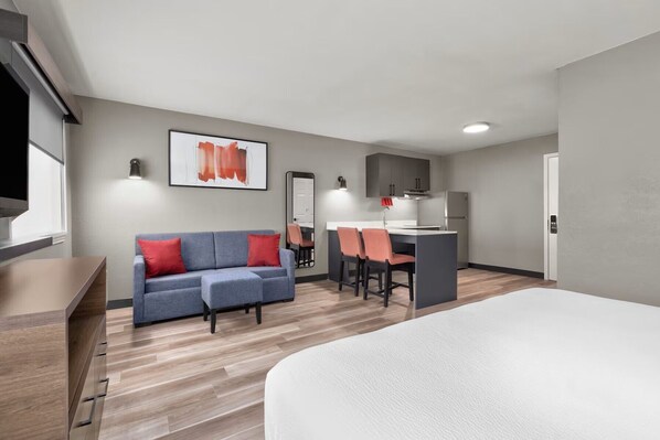 Una habitación de hotel con cama, sofá con cojines rojos, pequeña cocina con refrigerador y mesa de comedor con sillas.