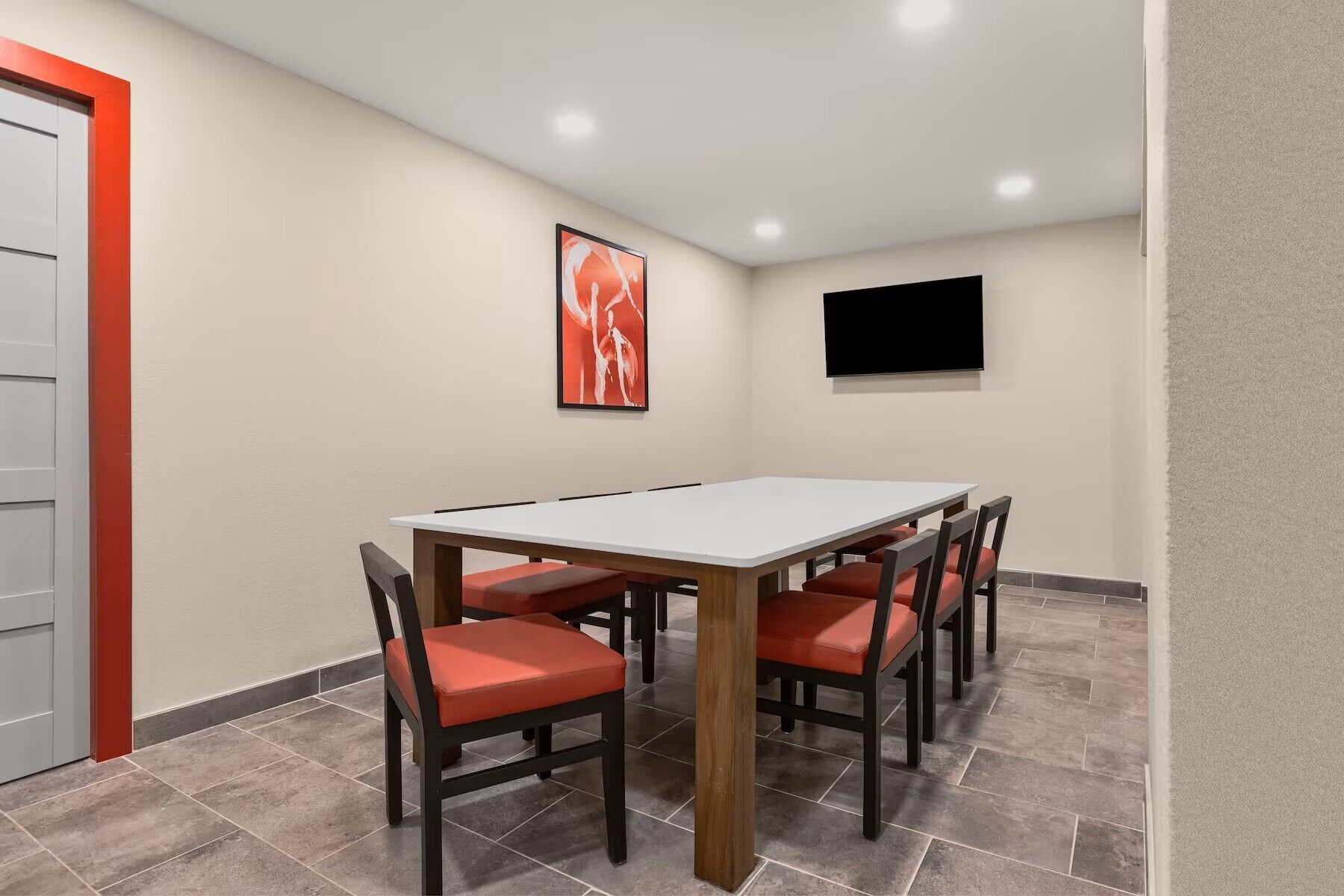 A conference room with a white table, red chairs, a framed picture, and a TV on the wall.