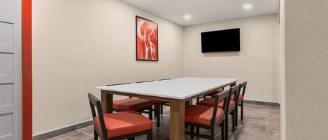 A conference room with a white table, red chairs, a framed picture, and a TV on the wall.