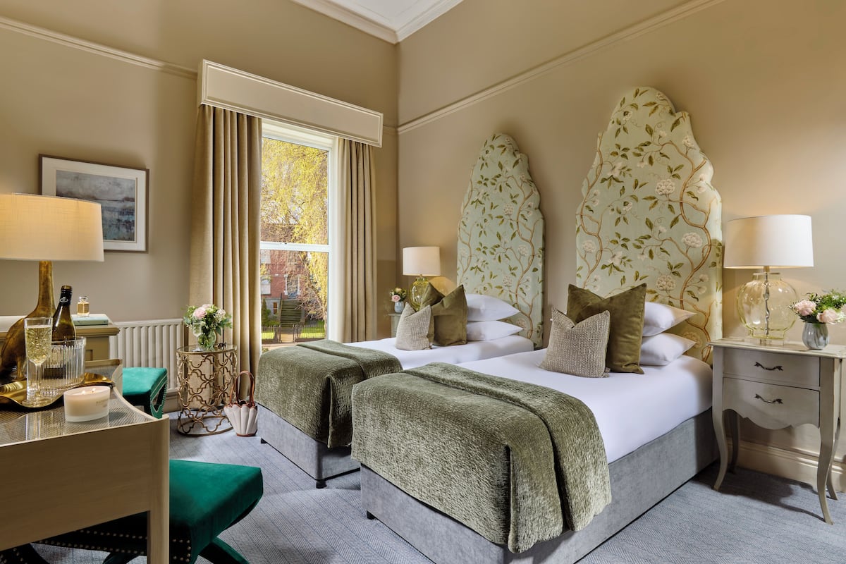 A double bed with a floral headboard, two bedside tables with lamps, a view of the outdoors, and a small table with a bottle and glasses.