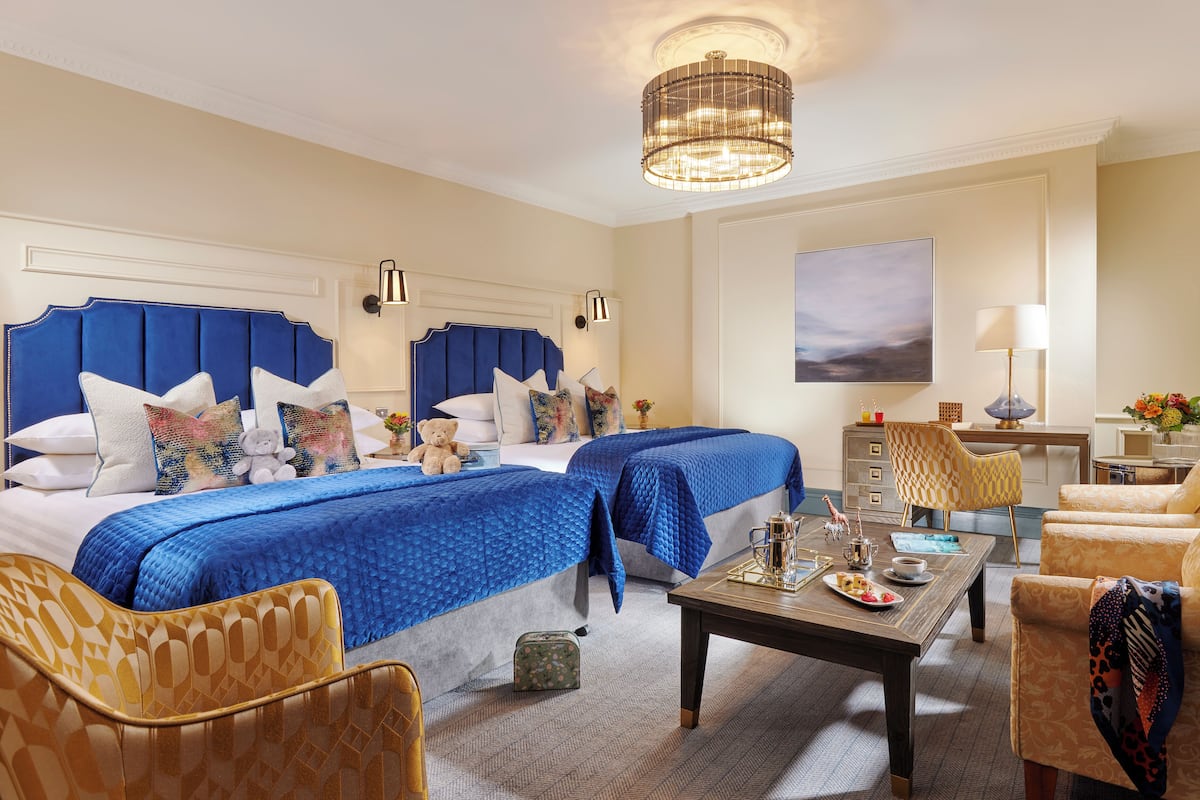 A hotel room with two beds, a dining table, a chair, and a lamp.