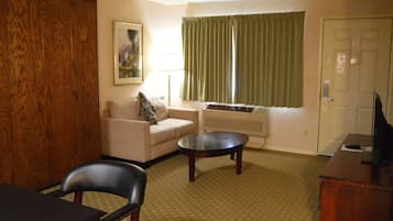 A hotel room with a wooden wardrobe, a sofa with a leopard print pillow, a round coffee table, a TV on a wooden stand, and a window with curtains.