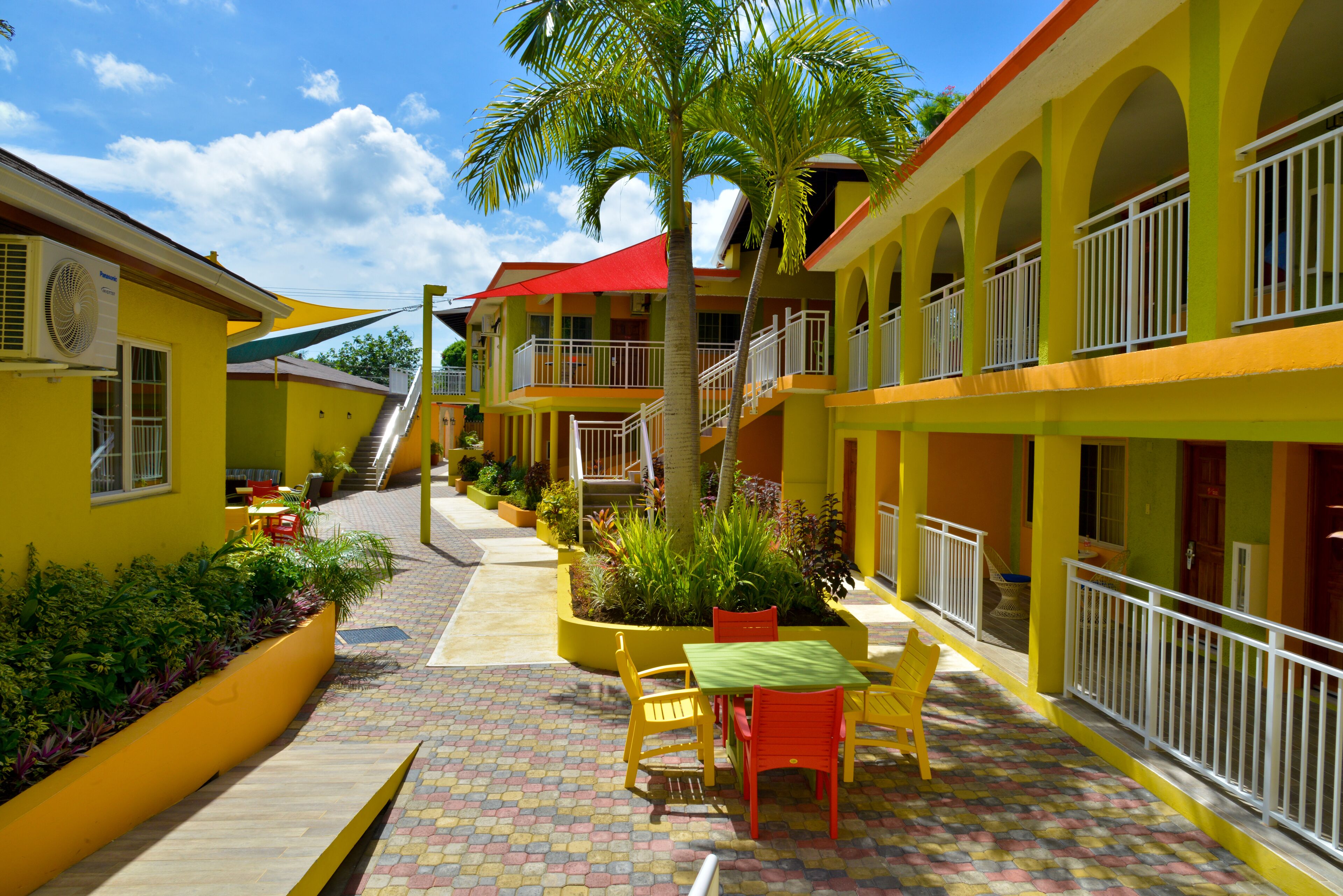 A hotel with a courtyard featuring a central seating area with chairs and a table, surrounded by plants and a palm tree. There are multiple balconies with railings, and a staircase leading to an upper level.