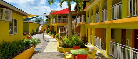 A hotel with a courtyard featuring a central seating area with chairs and a table, surrounded by plants and a palm tree. There are multiple balconies with railings, and a staircase leading to an upper level.