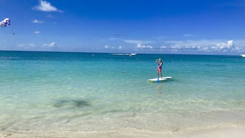 A beach with clear turquoise water, a person paddleboarding, and a boat in the distance.