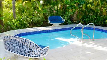 Una piscina con borde de azulejos azules, rodeada de exuberante vegetación y dos sillas de mimbre blanco.