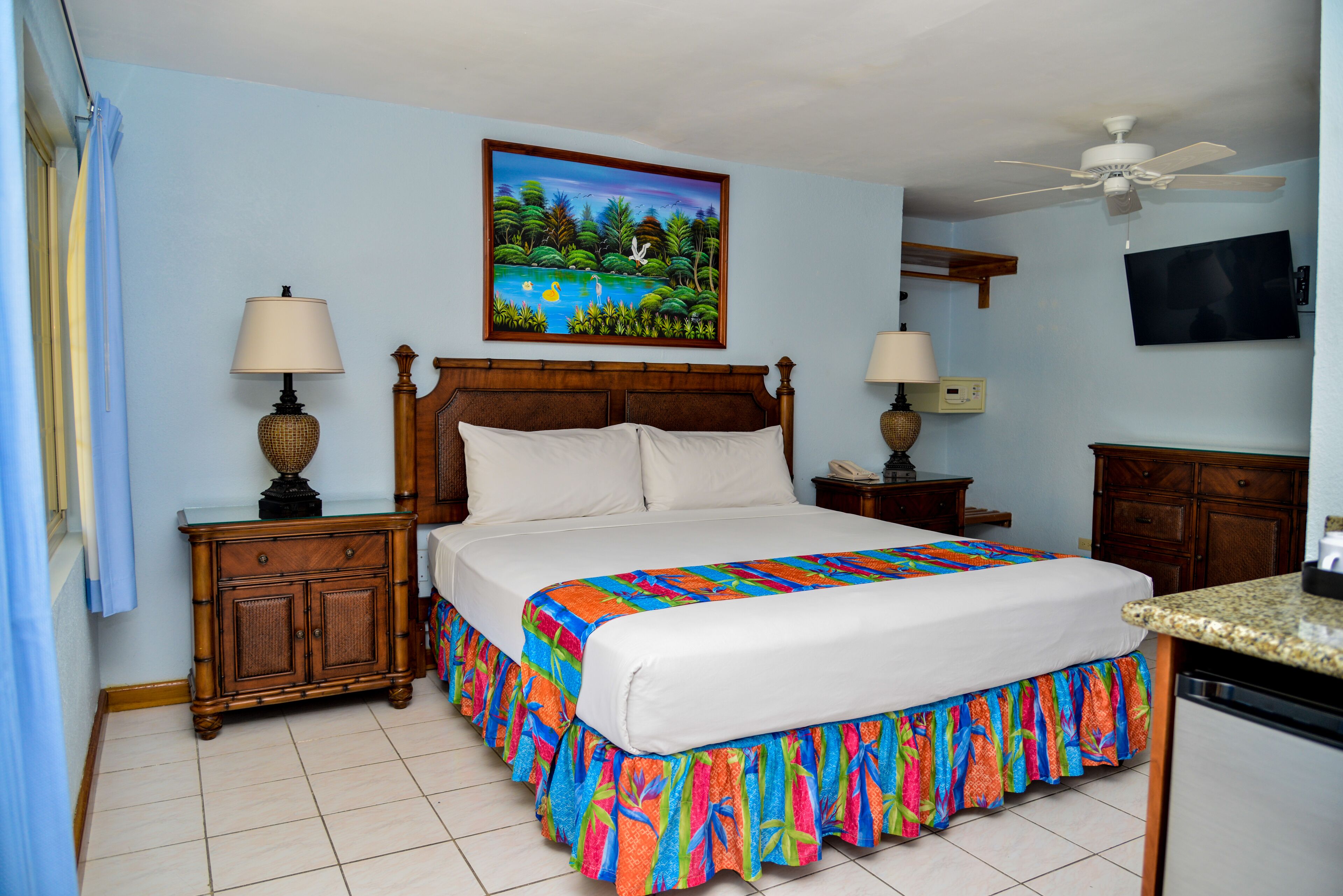 A bedroom with a bed, two bedside tables, a ceiling fan, a TV, and a painting on the wall.