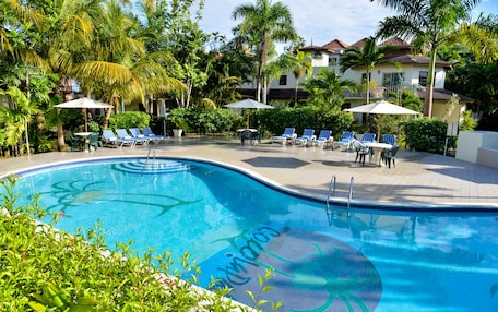 A swimming pool with lounge chairs and umbrellas, surrounded by palm trees and a building with a tiled roof.