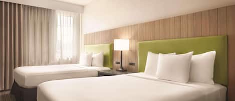 Una habitación de hotel con dos camas, un ventanal con cortinas, un cabecero de madera y una lámpara de noche.