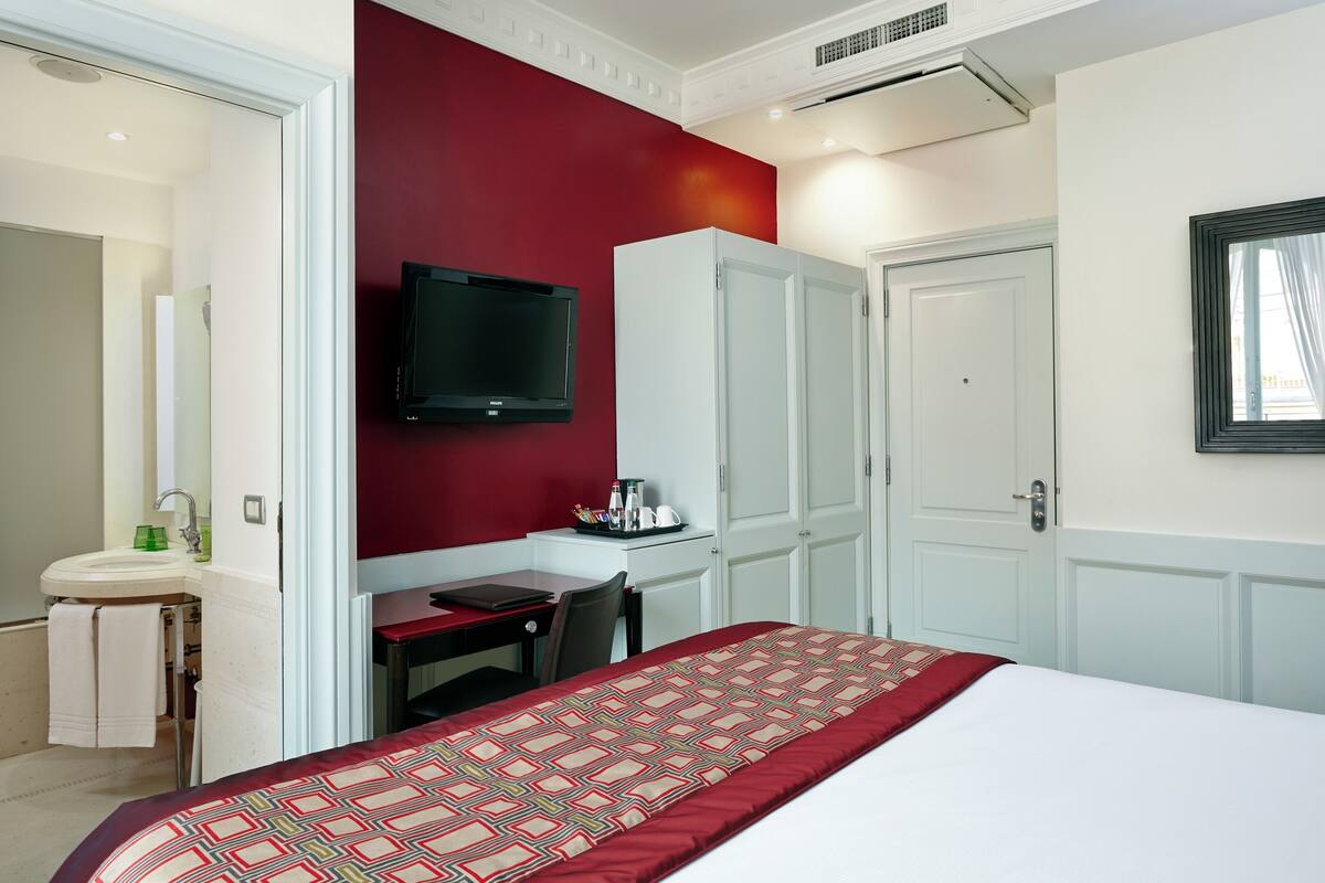 A hotel room with a bed, a desk with a chair, a TV mounted on the wall, and a wardrobe.