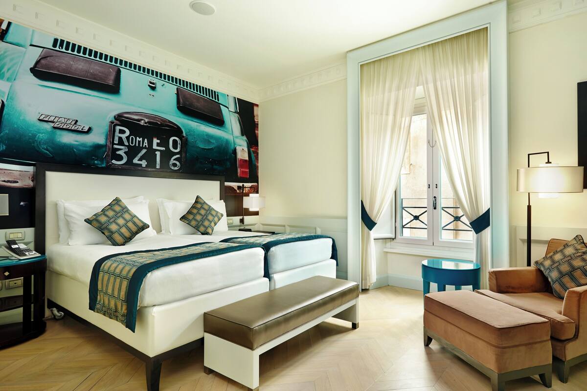 A hotel room with a bed, bedside table, bench, chair, and a large window with curtains.