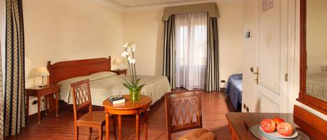 A hotel room with a bed, a desk with a chair, a small table with a plant, and a dresser with apples on it.