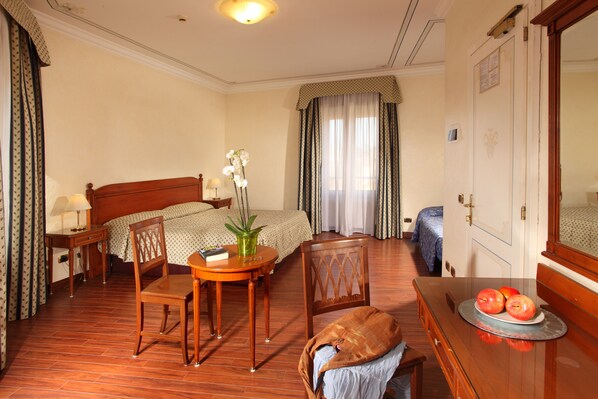 Une chambre d’hôtel avec un lit, un bureau et une chaise, une petite table avec une plante, et une commode sur laquelle sont posées des pommes.