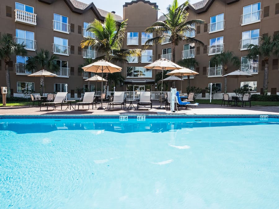 A swimming pool with lounge chairs and umbrellas, surrounded by a multi-story building with balconies and palm trees.