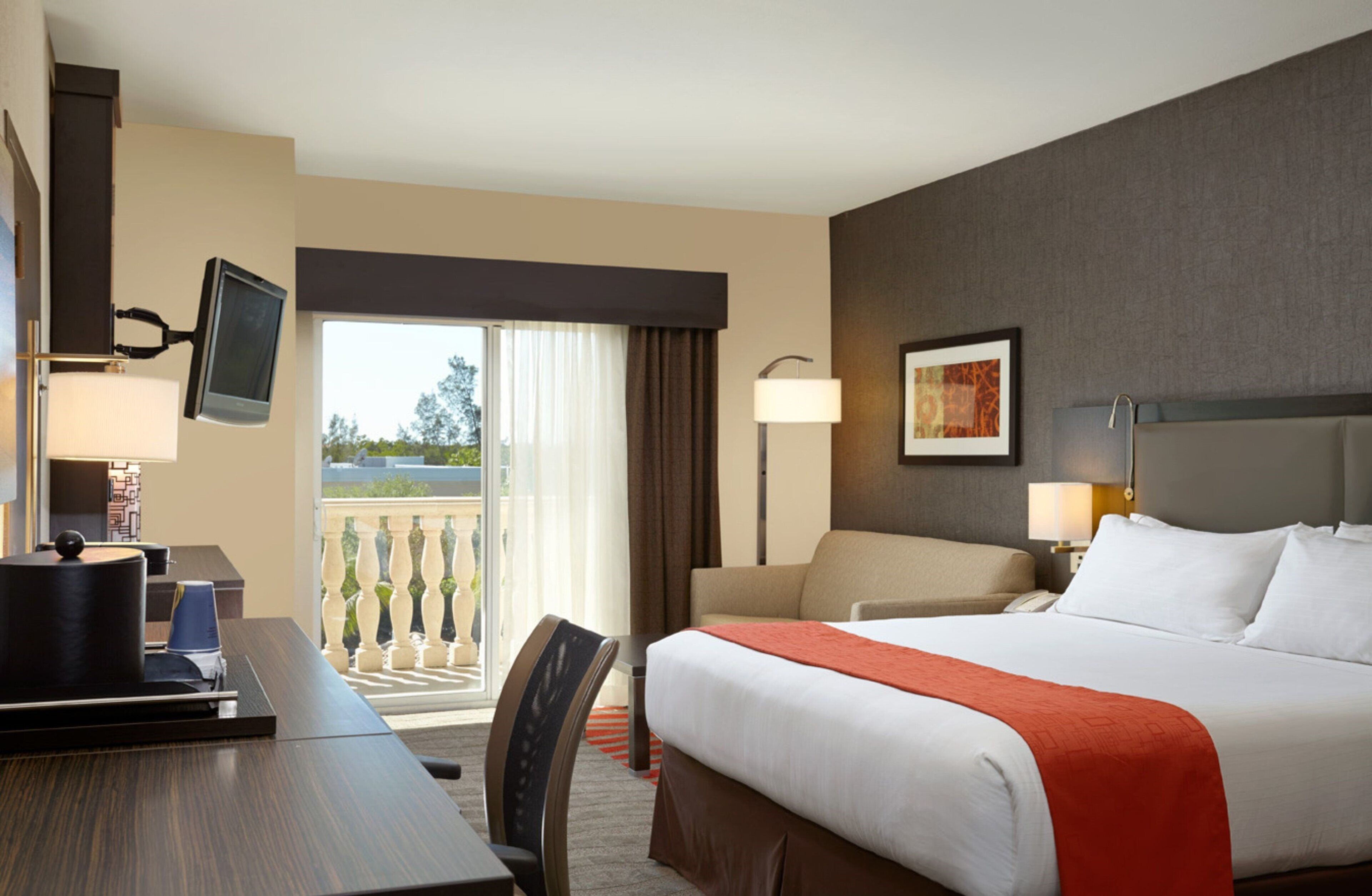 A hotel room with a bed, a desk, a chair, a TV mounted on the wall, a balcony with a view, and a painting on the wall.