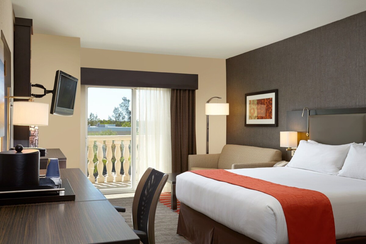 A hotel room with a bed, a desk, a chair, a TV mounted on the wall, a balcony with a view, and a painting on the wall.