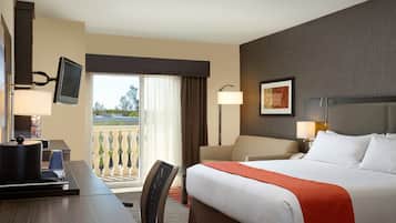 Habitación de hotel con cama, escritorio, silla, televisor montado en la pared, balcón con vistas y un cuadro en la pared.