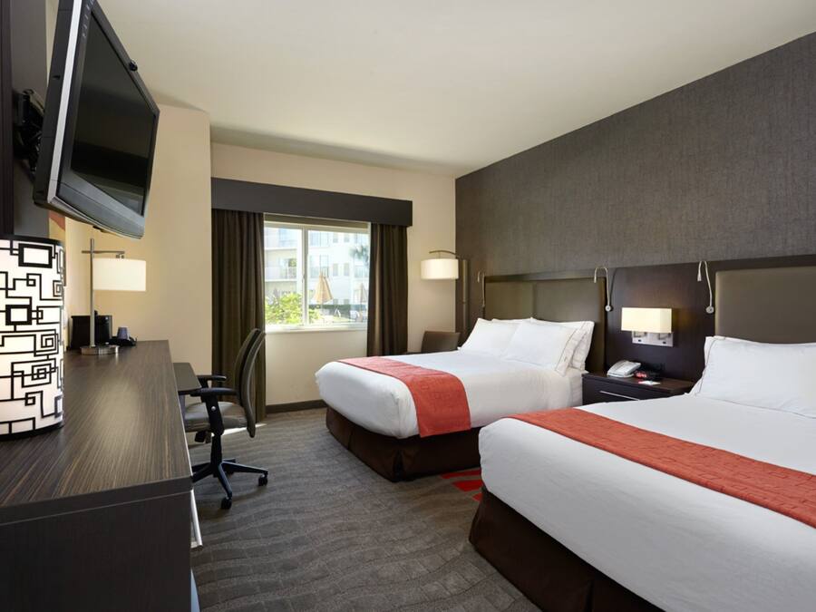 A hotel room with two beds, a desk with a TV, a lamp, and a view of the outside.