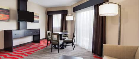 Habitación de hotel con televisión de pantalla plana, escritorio, mesa de comedor con sillas, sofá y un ventanal con cortinas.