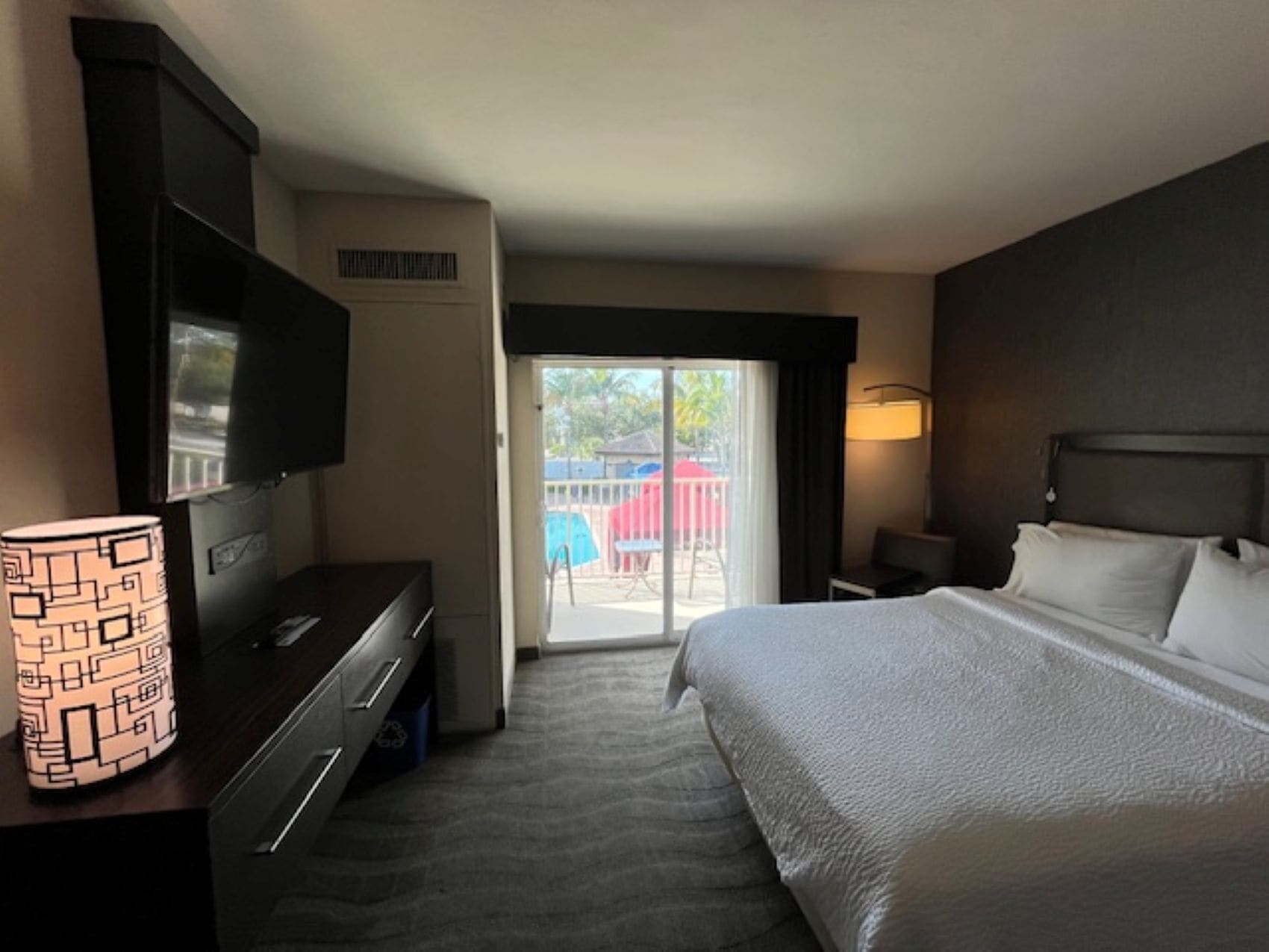 A hotel room with a bed, a TV, a desk, a chair, a lamp, and a sliding door to the balcony.