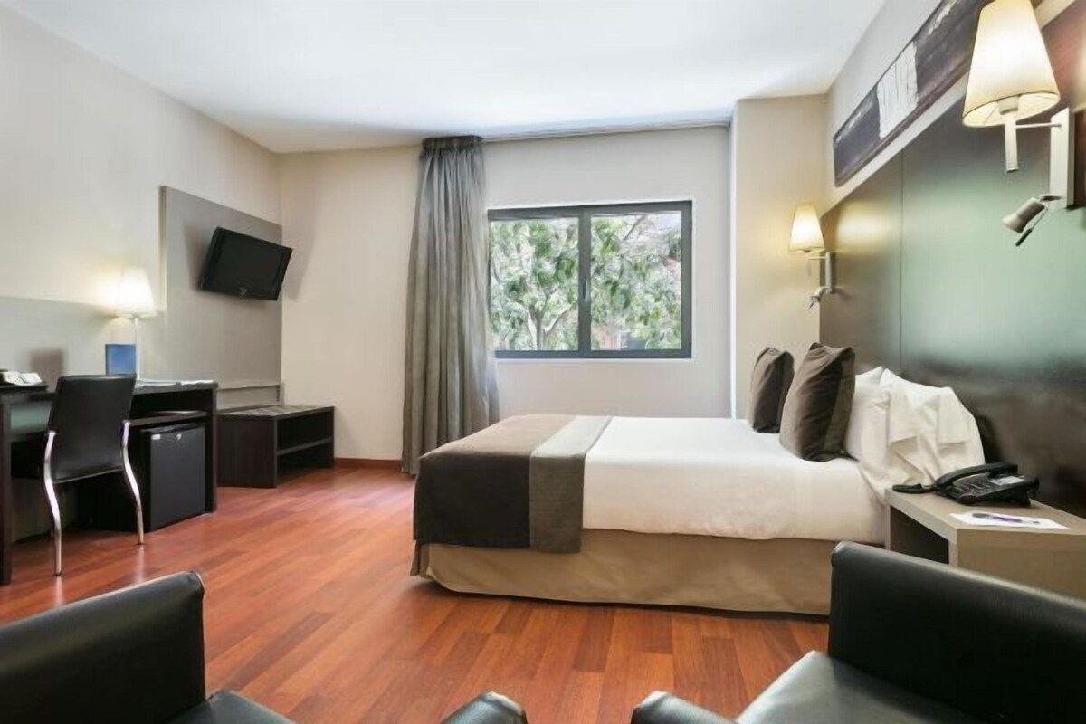 Una habitación de hotel con cama, escritorio, silla y televisor montado en la pared.