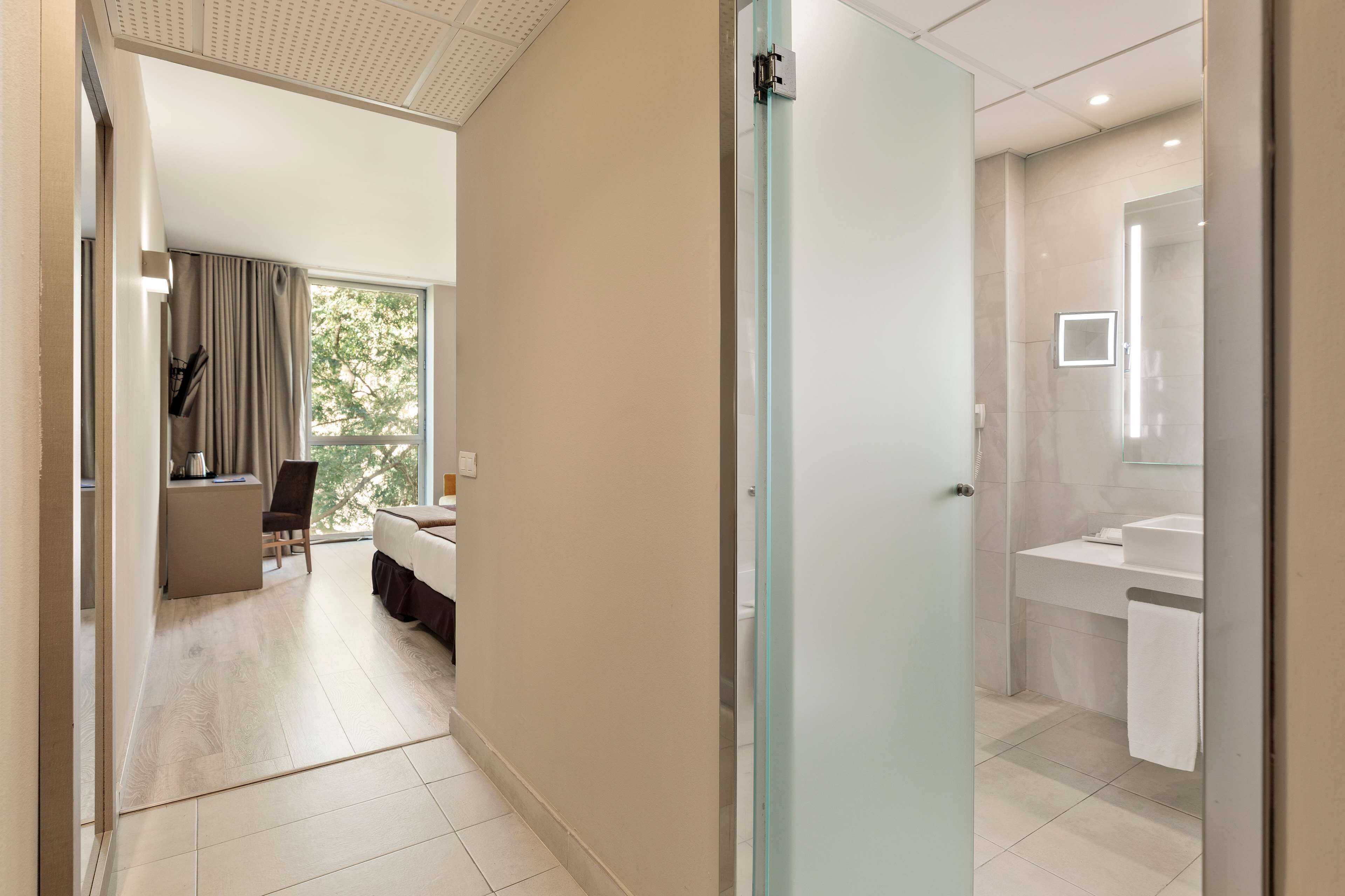 A hotel room with a bed, a small desk with a chair, a window with curtains, and a bathroom with a sink and mirror.