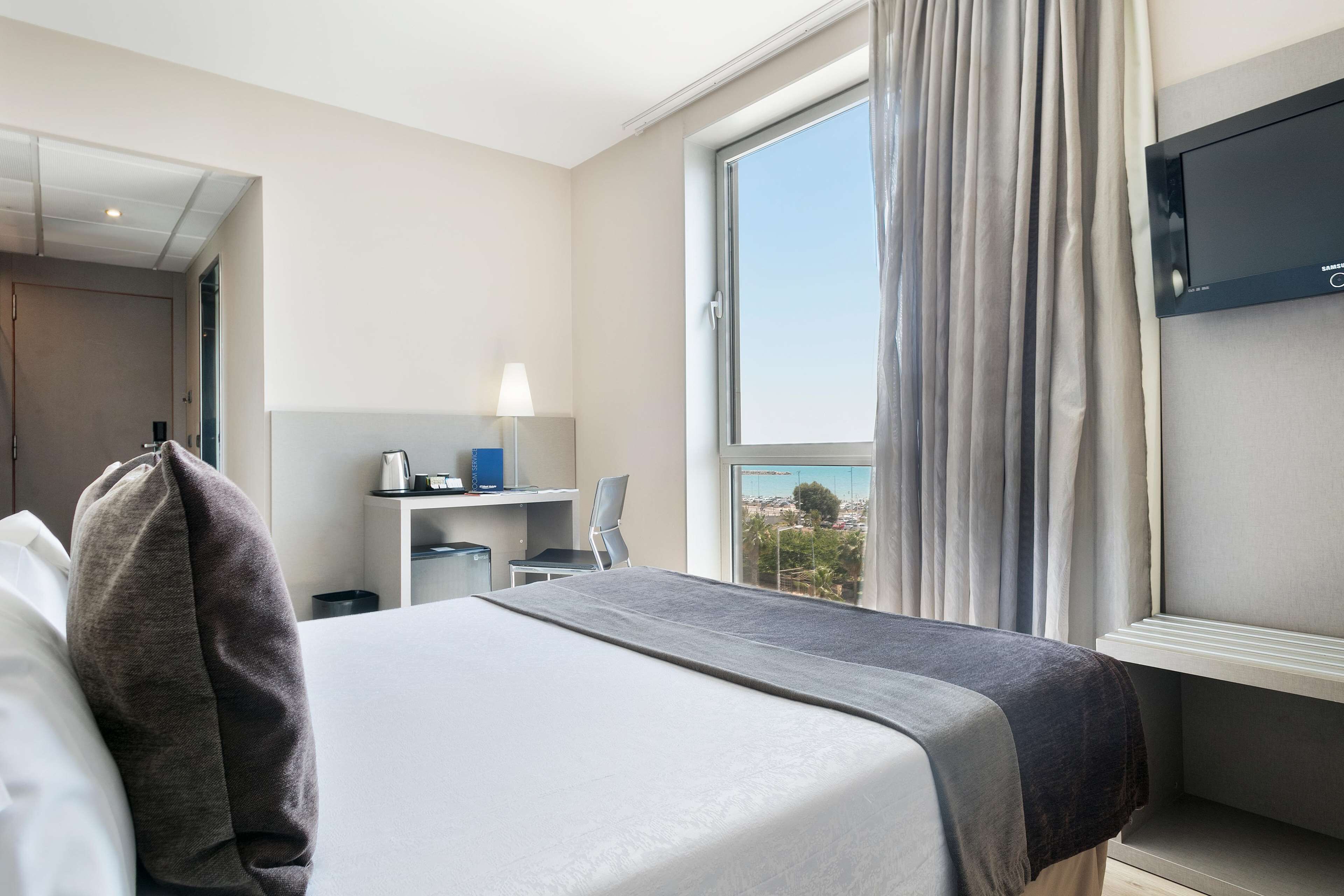 Una habitación de hotel con cama, escritorio, silla, televisor y ventana con cortinas.