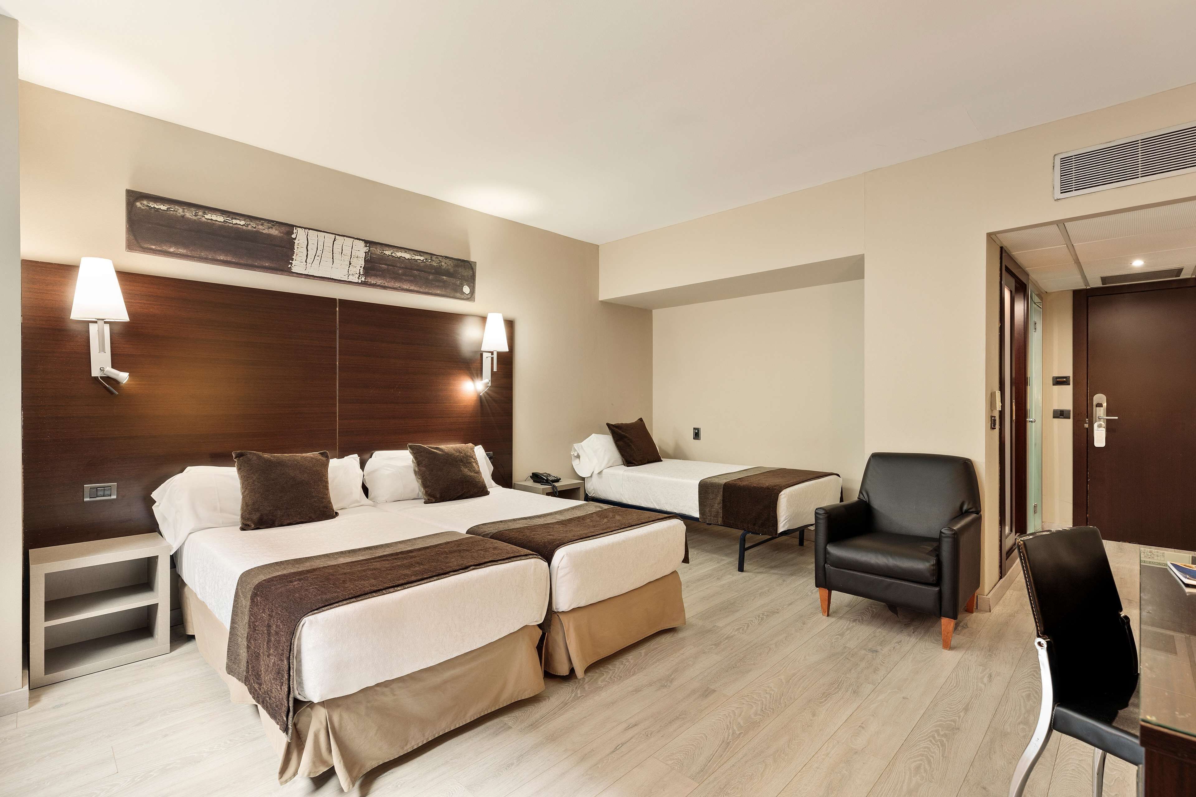 A hotel room with two beds, a desk, a chair, and a lamp.