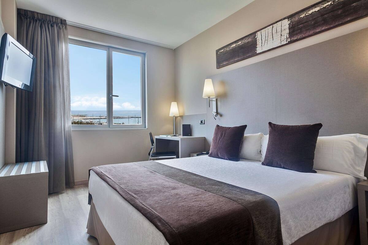 A hotel room with a large bed, a desk, a chair, a TV, and a window with a view of the sea.
