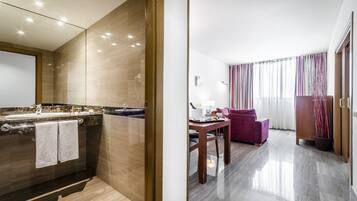Habitación de hotel moderna con baño que incluye un lavamanos, espejo y perchero. La habitación tiene piso de madera, una mesa de comedor con sillas y un ventanal con cortinas.