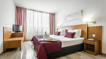 A hotel room with a bed, a TV, a bedside table, a lamp, and a window with curtains.