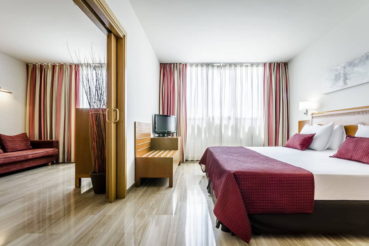 A hotel room with a bed, a TV, a sofa, and a wooden bench.