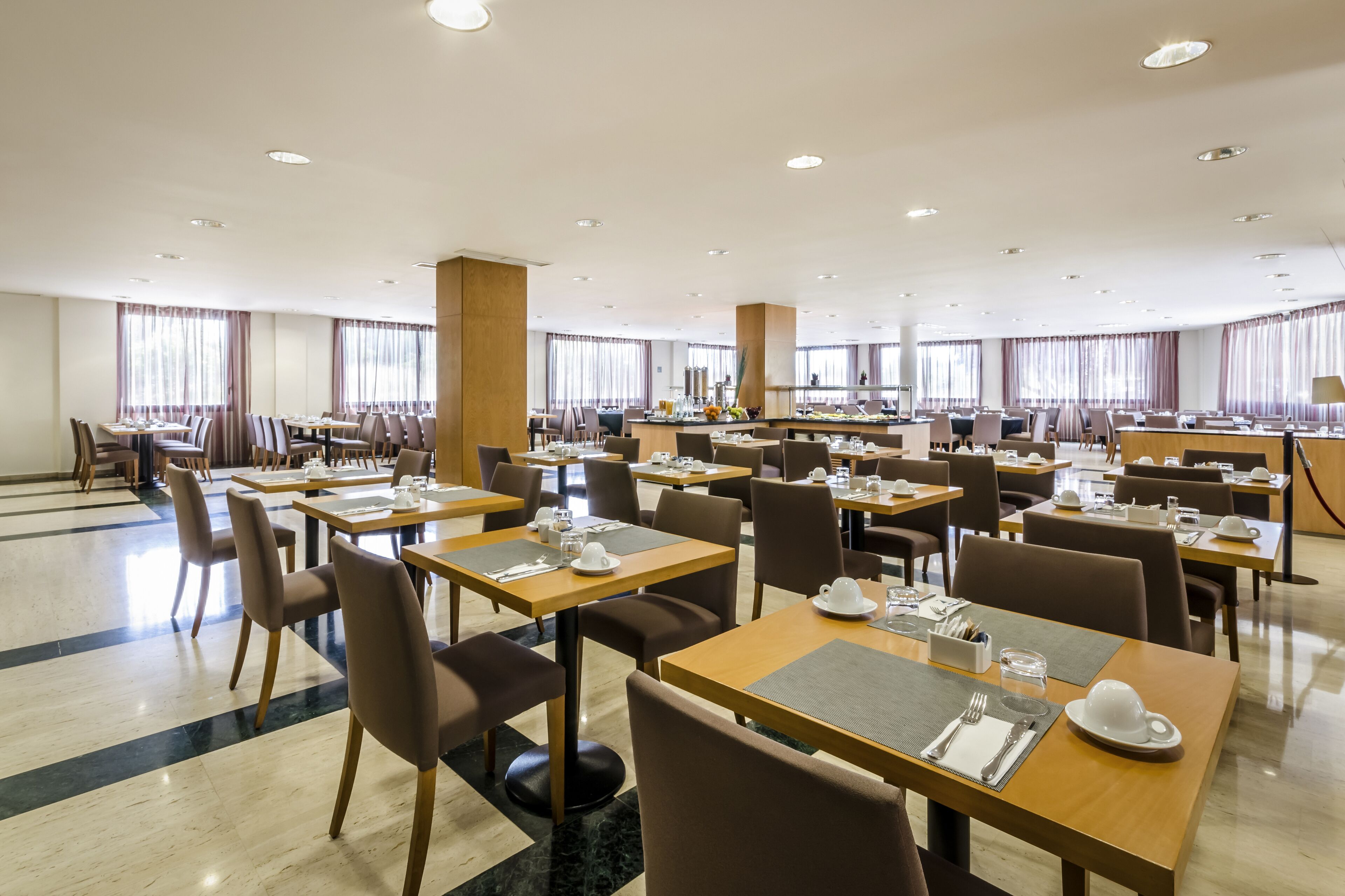 Een ruime hotel eetruimte met meerdere tafels en stoelen, grote ramen met gordijnen, en een schoon, modern design.
