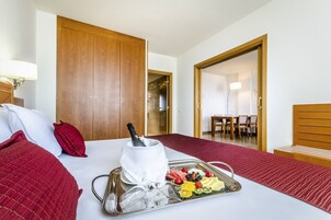 Habitación de hotel con una cama, una bandeja con frutas y una botella, y vista a un comedor.