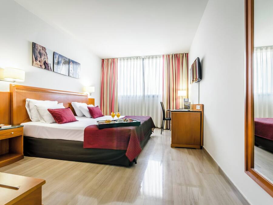 Quarto de hotel com cama, mesas de cabeceira, escrivaninha, cadeira e televisão.