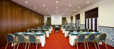 Un salón de conferencias con filas de mesas y sillas, una alfombra roja, paneles de madera en una pared y un revoque de baldosas en otra.