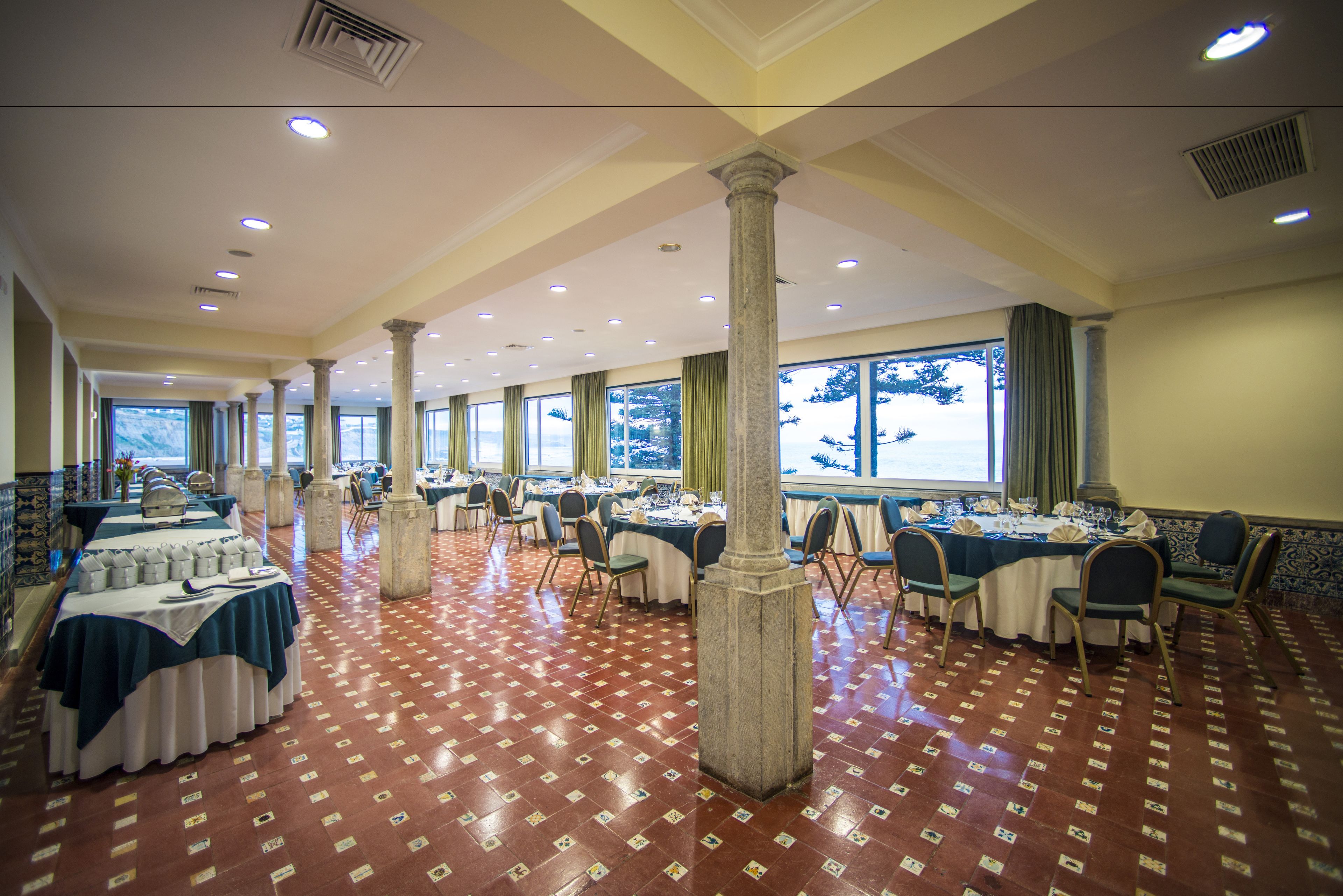 A spacious banquet hall with multiple round tables, high ceilings, and large windows.