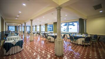 A spacious banquet hall with multiple round tables, high ceilings, and large windows.